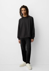 Mlada osoba s dredovima nosi labavu crnu majicu s dugim rukavima, crne traperice i bijele tenisice, stoji naslonjena na bijeli zid.