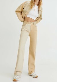 Femme portant un pantalon bicolore beige et crème, un crop top blanc, une veste beige et des baskets blanches, se tenant devant un fond clair.