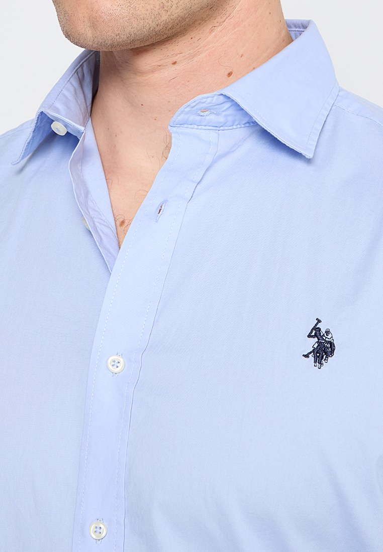 Camicia azzurra chiara con colletto classico, caratterizzata da bottoni bianchi e un piccolo logo ricamato scuro sul petto. Tessuto dalla consistenza liscia.