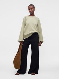 Femme portant un sweat-shirt oversize vert clair, un pantalon large noir, des tongs et tenant un grand sac fourre-tout marron sur fond blanc.