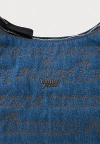 Tecido de denim com um tom azul, apresentando costura castanha em contraste. Logótipo "tommy jeans" bordado em detalhe metálico prateado perto do decote.