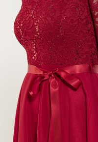 Robe en dentelle rouge avec un corsage ajusté orné de sequins et une jupe fluide. Comprend un ruban en satin à la taille pour plus de détails.