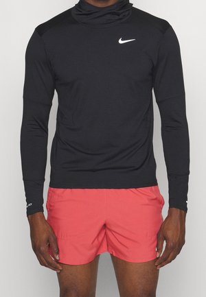 Mężczyzna ubrany w czarną, długorękawową koszulkę sportową Nike z białym logo oraz koralowoczerwone spodenki sportowe, stojący na jednolitym tle.
