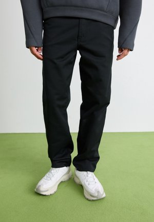 Personne portant un pantalon noir, un haut à manches longues foncé et des baskets blanches, debout sur un tapis vert avec un fond blanc.