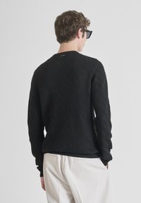 Zwarte trui met een geprofileerd visgraatpatroon, ronde hals en lange mouwen. Geribbelde manchetten en onderkant. Draagt over lichtgekleurde broek.