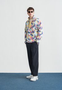 Νεαρός άνδρας που φοράει πολύχρωμο πουλόβερ με σχέδια, μαύρο παντελόνι, λευκά αθλητικά παπουτσάκια και γυαλιά ηλίου, στέκεται σε μπλε χαλί με λευκό φόντο.