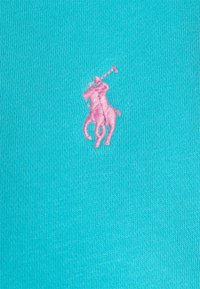 Tyrkysová látka s ribovanou strukturou, na které se nachází výrazné růžové vyšívané logo hráče póla.