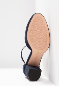 Chaussure à talon haut en daim bleu marine avec bout ouvert et bride autour de la cheville. La semelle extérieure est marron clair avec un motif texturé pour l'adhérence.