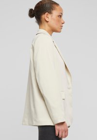 Blazer crema con revers a catena, maniche lunghe e tasche frontali. Tessuto morbido con una consistenza liscia, silhouette non strutturata. Pantaloni grigi indossati sotto.