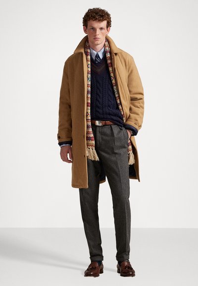 Tan wolcoat over een marineblauwe kabeltrui, grijze broek en bruin leren schoenen. Een multicolore franjesjaal voegt detail toe.