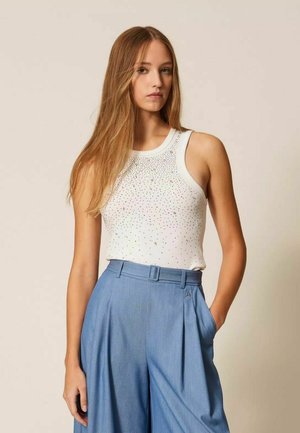 Giovane donna con lunghi capelli biondi che indossa un top bianco senza maniche con piccoli dettagli e pantaloni larghi a vita alta di colore azzurro chiaro.