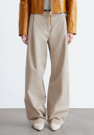 Person iført beige bukser med vide ben, hvide spidse sko, grå top og en blank brun jakke med ringe på fingrene.