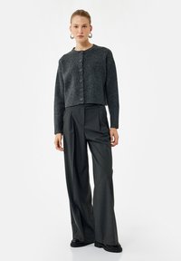 Kvinde iført en mørkegrå knap-sluttet cardigan, brede charcoal bukser, sorte sko og store hoops øreringe, stående mod en hvid baggrund.