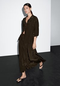 Olivgrön satinblus med lös passform, i kombination med en matchande svävande kjol. Modellen bär svarta gummiflip-flops. Minimalistisk bakgrund.