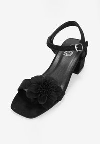 Sandal de ante negro con punta cuadrada, tira en el tobillo con hebilla plateada y acento de flor en la parte frontal. Diseño de tacón bajo en bloque.