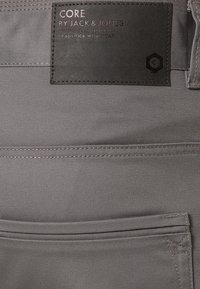 Pantalon de travail gris avec une étiquette en cuir noir embossée de texte. Le tissu est lisse avec une texture subtile et des détails de coutures.