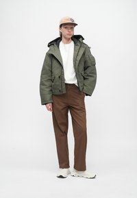 Olīvzaļš pufīgais jaka ar kapuci, valkāta virs baltas džemperes, kopā ar brūniem biksēm un baltām kedām. Tans cepure ar logo.