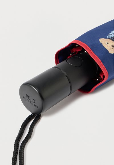 Λαβή μαύρης ομπρέλας με κυλινδρικό σχήμα, διαθέτει κόκκινο και μπλε υφασμάτινο κάλυμμα με σχέδιο αρκούδας, περιλαμβάνει πλεκτό μαύρο λουρί για μεταφορά.