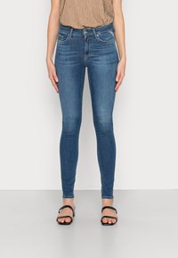 Jeans skinny de denim en azul medio, con una cintura alta, diseño de cinco bolsillos y una textura suave. Llevados con sandalias planas negras.