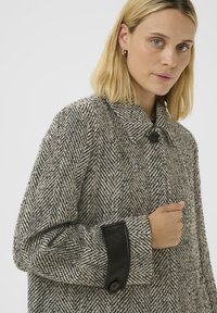 Femme blonde portant un manteau à motif chevrons noir et blanc avec un col, un bouton noir unique et des poignets en cuir avec un détail de bouton.