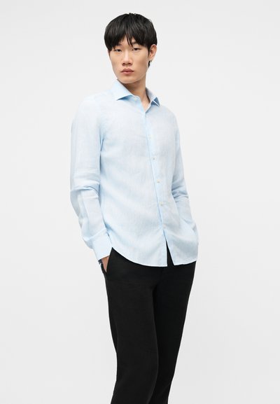 Νεαρός άνδρας στέκεται με τα χέρια στις τσέπες, φοράει ένα ανοιχτό μπλε πουκάμισο με κουμπιά και μαύρο παντελόνι, σε ένα απλό λευκό φόντο.