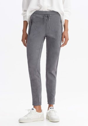 Person iført grå slim fit-bukser med lynlås-lommer foran og hvide sneakers, stående mod en hvid baggrund.