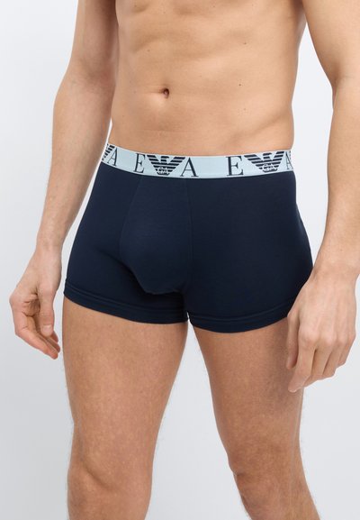 Ανδρικός κορμός που φοράει σκούρο μπλε μποξεράκια με ανοιχτό μπλε ζώνη μέσης που φέρει το λογότυπο EA και το έμβλημα αετού, στέκεται μπροστά σε απλό λευκό φόντο.