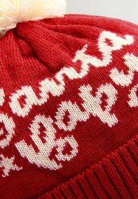 Bonnet tricoté rouge avec un texte blanc. Le dessus est décoré d'un pompon en crème duveteux. Le texte apparaît dans une police cursive, ludique.