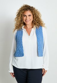 Chaleco acolchado azul con un patrón texturizado, llevado sobre una camisa blanca de botones. Presenta cortes laterales y bordes redondeados, combinado con pantalones oscuros.
