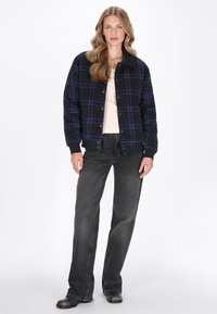 Knäppt jacka i marinblå och svart rutigt mönster, tillverkad av textil med struktur. Kombineras med en ljus topp och mörkgrå jeans, samt svarta skor.