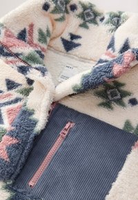 Veste polaire douce crème avec des motifs géométriques roses, verts et bleus, avec une poche poitrine en velours côtelé bleu et une fermeture éclair rose.