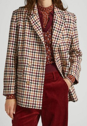 Kvinna i en flerfärgad houndstooth dubbelknäppt blazer, mönstrad marsala-halsvärmare, bordeaux geometrisk tröja och röda kordbyxor.