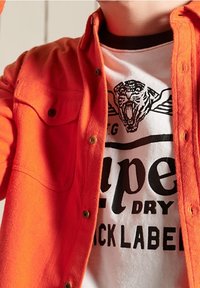 Oranssi nappipaita valkoisen T-paidan päällä, jossa on tiikerin kuva ja mustaa tekstiä. Paidassa on sile�ä textura ja näkyvät napit.