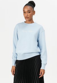 Light blue sweatshirt featuring a crew neckline and ribbed cuffs. "Tommy Hilfiger" is embroidered in white. Paired with a black pleated skirt.