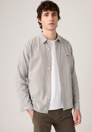 Junger Mann mit lockigem dunklem Haar, der ein hellgraues Hemd über einem weißen T-Shirt und braunen Hosen trägt, steht vor einem schlichten Hintergrund.