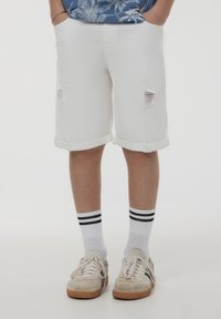 Shorts di denim bianco con orli arrotolati e dettagli consumati, abbinati a calzini bianchi con strisce nere e sneakers chiare.