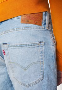 Jean Levi's bleu clair avec poche arrière visible, étiquette rouge Levi's et patch en cuir indiquant la taille W31 L porté par une personne portant un haut orange.