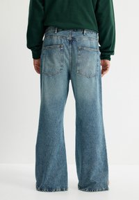 Jeans de tiro ancho en azul claro, hechos de denim, con un lavado desgastado, bolsillos traseros de parche y un detalle de dobladillo deshilachado.
