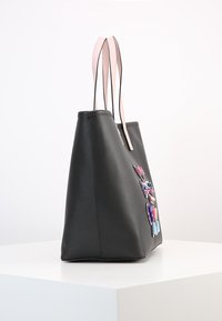 Svart tote-väska med ljusrosa handtag, prydd med färgglada emaljknappar, uppvisad på en vit pelare mot en enfärgad bakgrund.