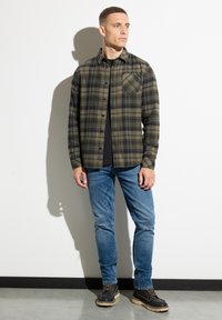 Camisa de cuadros en verde y negro con un bolsillo en el pecho, combinada con jeans azules y botas marrones. Líneas limpias y ajuste informal.