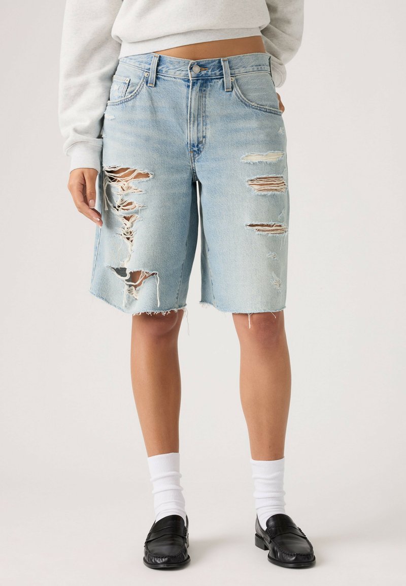 Person wearing light blue ripped denim shorts, white socks, black loafers, and a light-colored cropped sweatshirt.