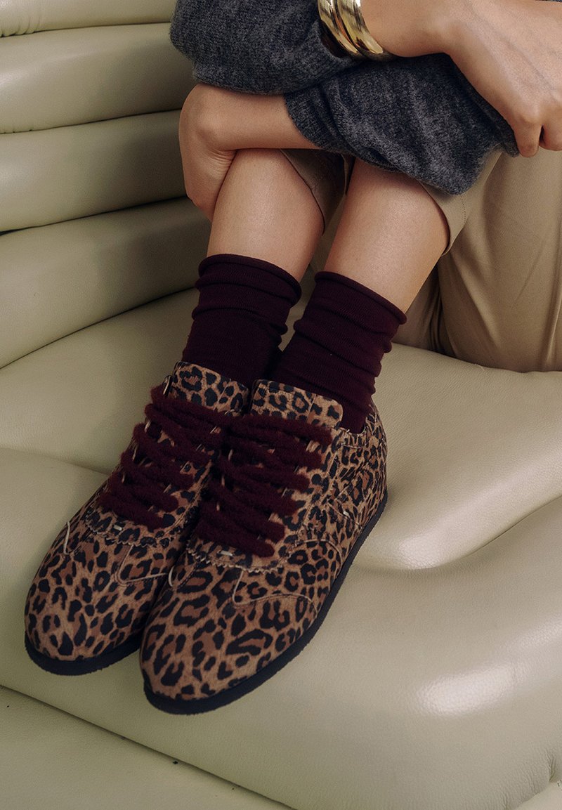 Baskets à imprimé léopard avec des lacets bordeaux, texture de tissu douce et semelles noires, associées à des chaussettes maroon, reposant sur une surface de couleur claire.