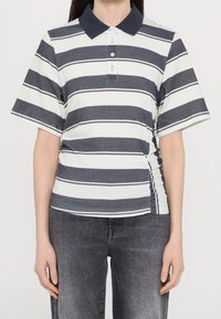 Personne portant un polo à manches courtes bleu marine et blanc à rayures, avec un jean noir, se tenant devant un fond blanc uni.