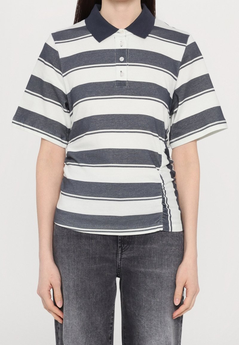 Personne portant un polo à manches courtes bleu marine et blanc à rayures, avec un jean noir, se tenant devant un fond blanc uni.