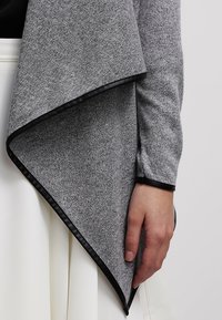 Personne portant un cardigan asymétrique texturé gris avec bordure noire sur une jupe plissée blanche, main détendue sur le côté.