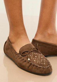Bruna mockasinläderskor med en dekorativ nitar design, en knuten rosett och en gummisula. Kanterna har intrikata sömnadsdetaljer.