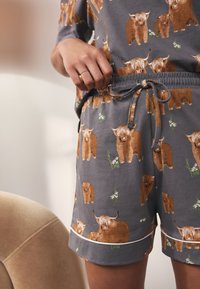 Grå bomuldsshorts med print af Highland-kvæg i brune og beige nuancer med grønne detaljer. Inkluderer elastisk talje og snøre.