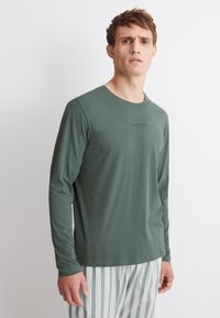 Olijfgroene katoenen shirt met lange mouwen en een ronde hals, voorzien van een subtiele logo-opdruk. Gekoeld met gestreepte, lichte onderkleding.