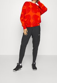 Sweat-shirt rouge avec un grand motif abstrait orange, associé à un pantalon de jogging noir fuselé. Chaussures de sport noires avec des accents blancs.