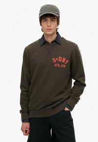 Olive zielenony sweter z grafiką "S&DRY" w kolorze pomarańczowym, długie rękawy, ściągacze na mankietach, nałożony na koszulę w kratę. Ubranie z czarnymi spodniami.